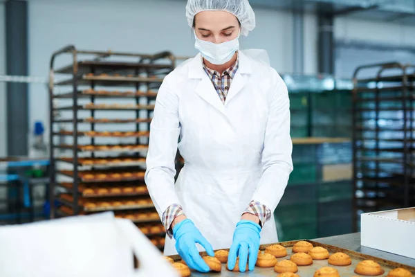 Confectionery factory worker in white coat taking delicious freshly baked pastry from tray.