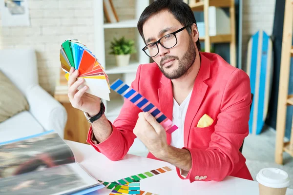 Confident young man working in atelier sitting at desk and choosing colors out of bright palettes.