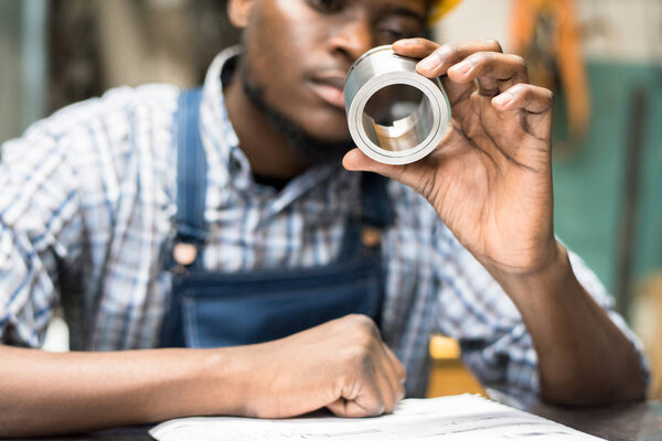 Close-up of concentrated young African-American engineer holding steel bushing and viewing it while checking detail in workshop