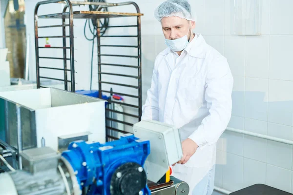 Confectionery factory employee in white coat standing and using machinery.