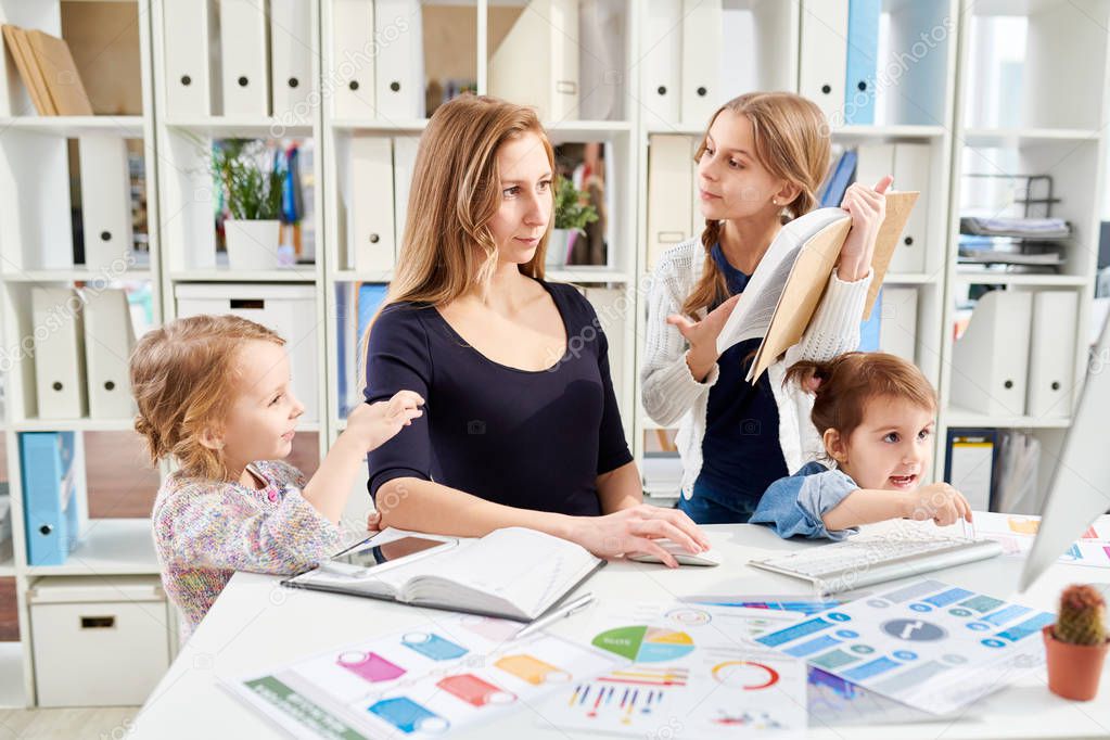 Young white collar worker sitting at desk and creating visual presentation for business partners while her little daughters trying to attract her attention, office interior on background
