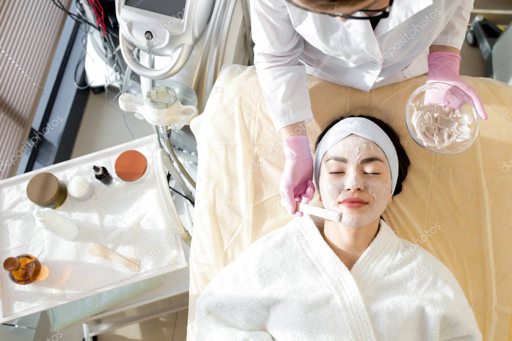 Directly above view of attractive young woman wearing bathrobe and headband lying on treatment table with closed eyes while receiving beauty treatment at spa salon