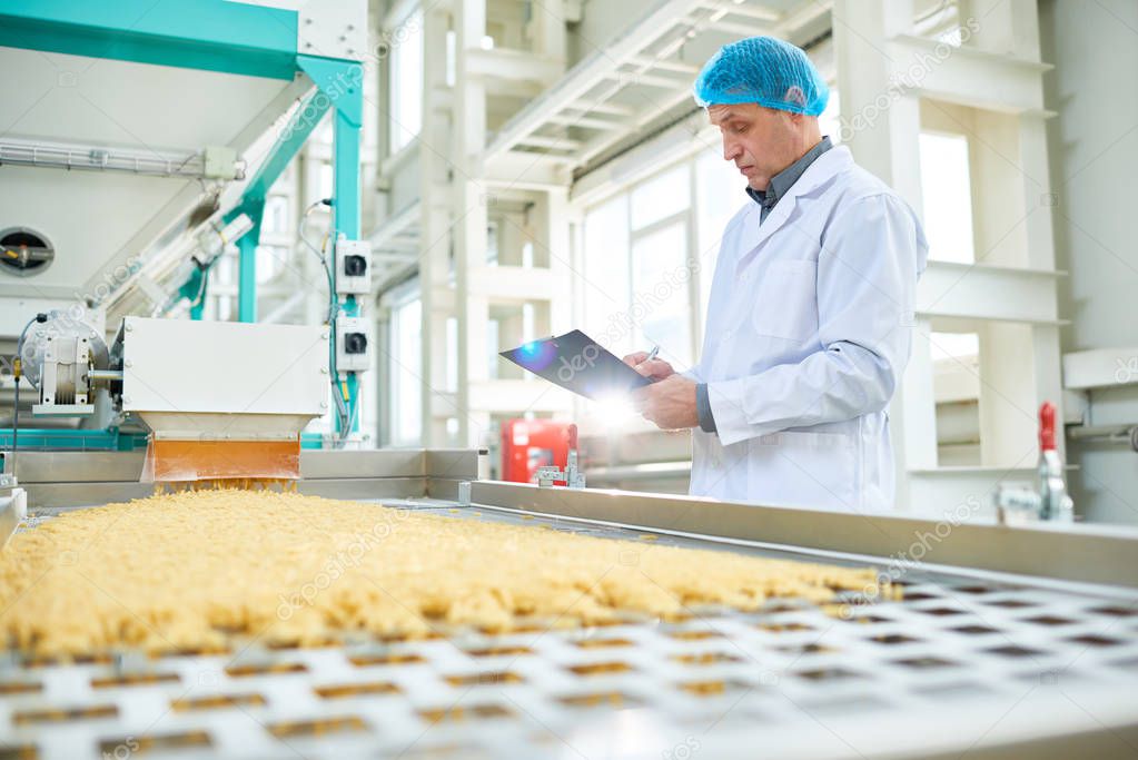 Side view portrait of senior factory worker  in food industry holding clipboard standing by conveyor belt, copy space