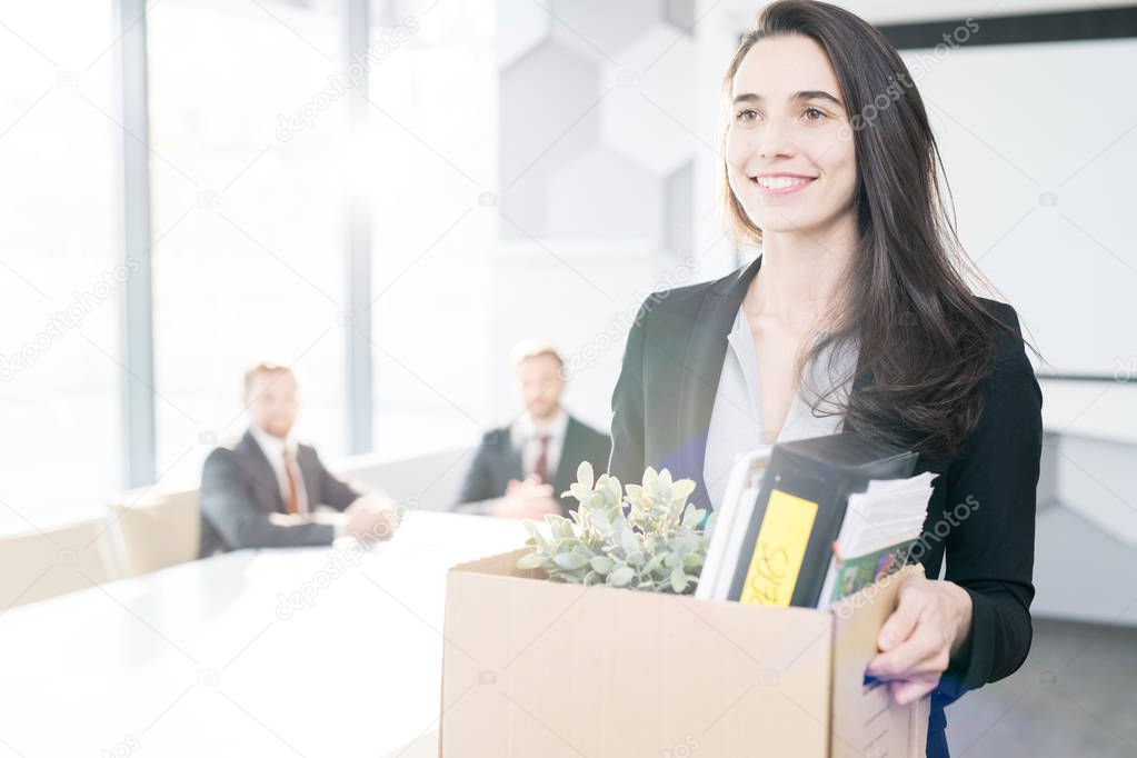 Waist up portrait of happy young businesswoman holding box of personal belongings  leaving office after quitting job