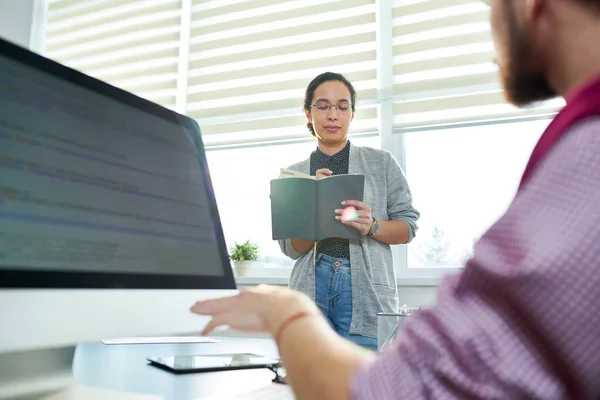 窓のそばに立って メモ帳で必要なデータを書く女性同僚中のオフィスにあるコンピューターで作業する人 — ストック写真
