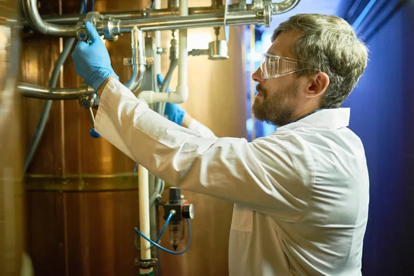 Koruyucu gözlük ve modern bira fabrikasında dururken silindirik bira olgunlaşma tankta basınç ayarlama beyaz önlük giymiş yakışıklı sakallı teknisyen