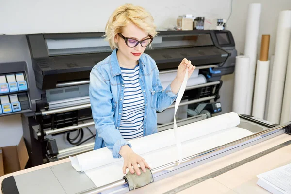 Beautiful young woman in glasses printing drafts on plotter during work in office.