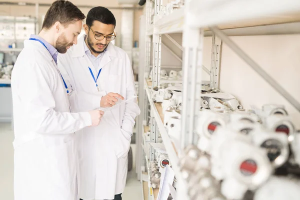 Multi-ethnic team of quality control engineers wearing lab coats studying document while standing at spacious warehouse of pressure transducer factory and carrying out inspection