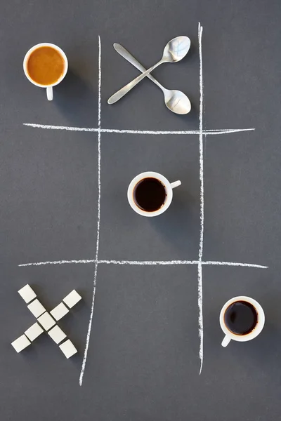 Top view of crossed spoons, coffee cups and sugar cubes on gray background with tic tac toe design, copy space