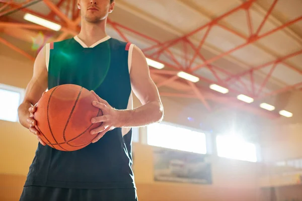 Crop man in basketball sportswear holding ball while standing on court in gym.