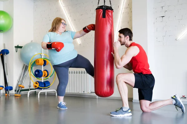 Full length portrait of cheerful obese woman hitting punching bag in gym during personal training with fitness instructor