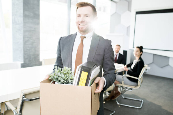 Waist up portrait of happy young man holding box of personal belongings leaving office after quitting job