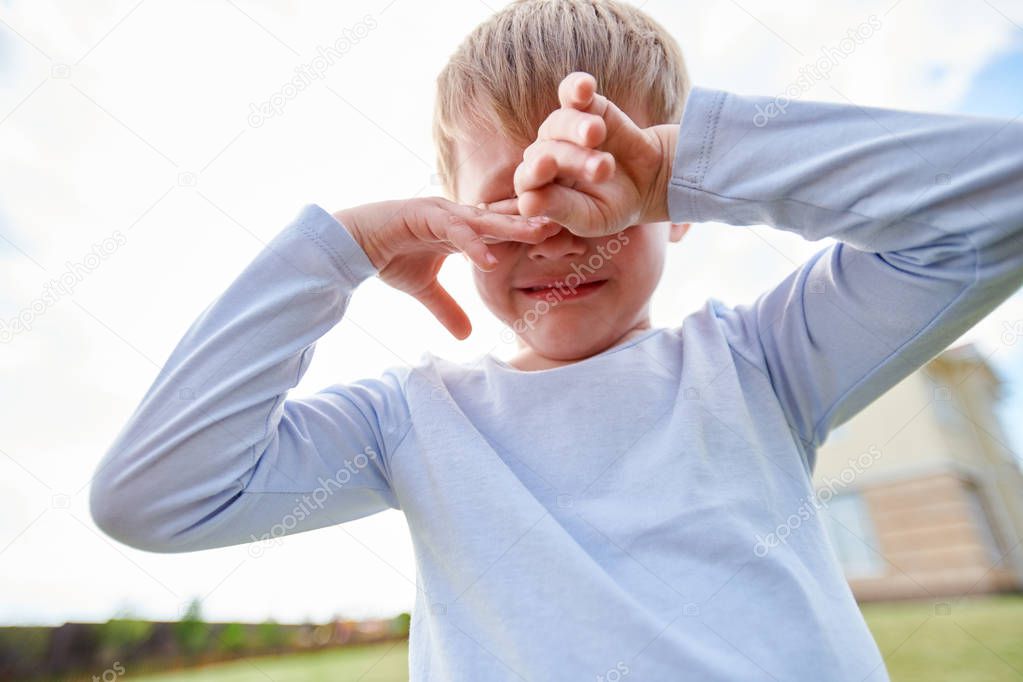 Low angle portrait of sad little boy crying hysterically outdoors, covering eyes with hands