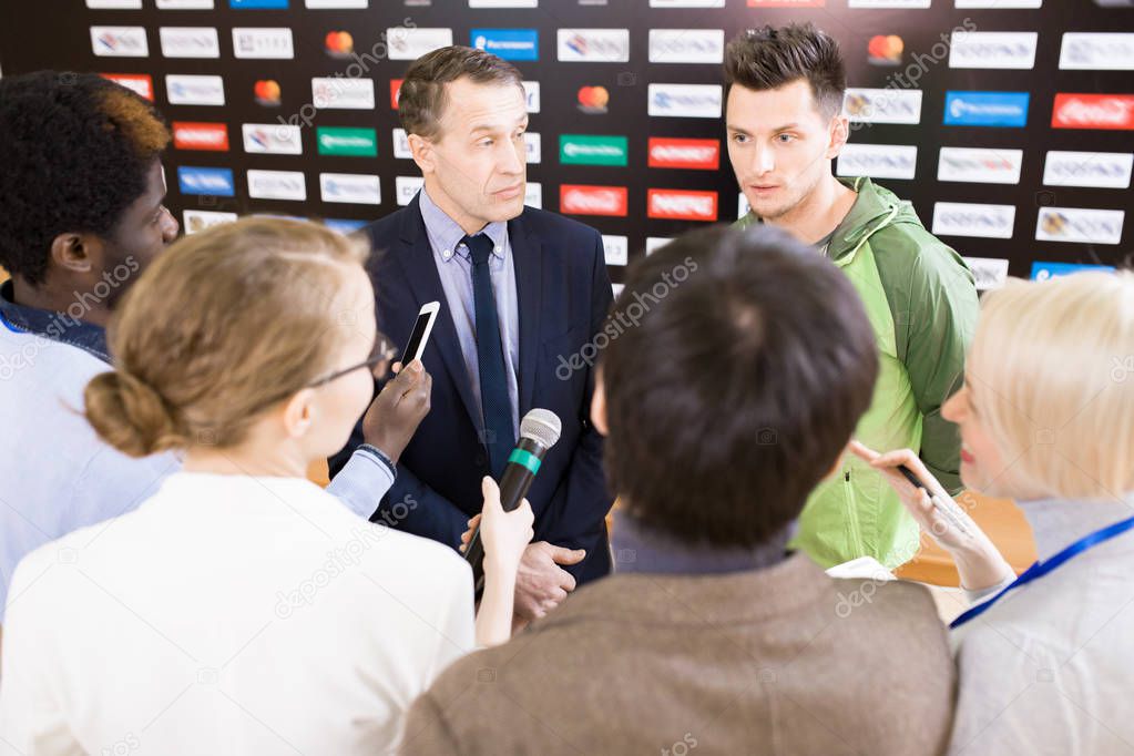 High angle view at international group of journalists interviewing sportsman at press conference, handing recording devices and microphones