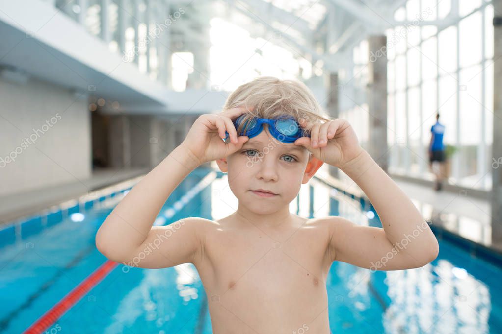 Calm cute boy with blond hair training in swimming pool: he putting on goggles and looking at camera