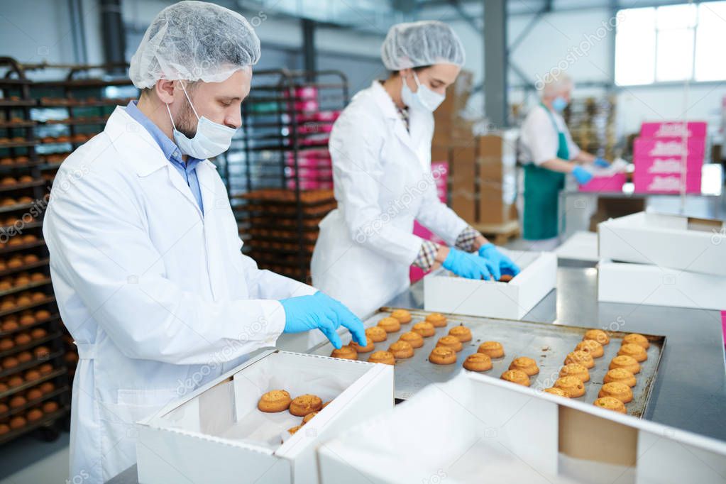 Confectionery factory employees in white coats collecting freshly baked pastry from tray and putting it into paper boxes.