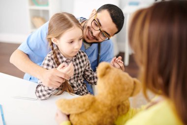 Modern klinik istişare sırasında annesiyle masa başında oturan sevimli küçük kız ile oynarken neşeli Ortadoğu çocuk doktoru portresi