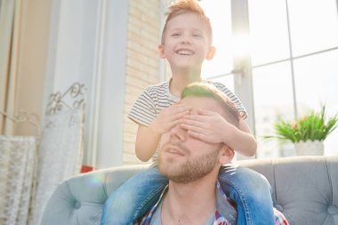 Portrait of cute little boy sitting on his fathers shoulders and covering his eyes while sitting on sofa against sunlit windows at home clipart