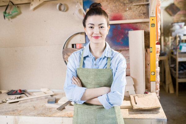 Waist up portrait of young female artisan posing in crafting shop standing with arms crossed, looking at camera and smiling