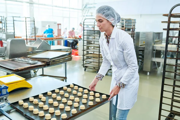 Confectionery factory worker in white coat standing and holding baking tray with uncooked pastry.