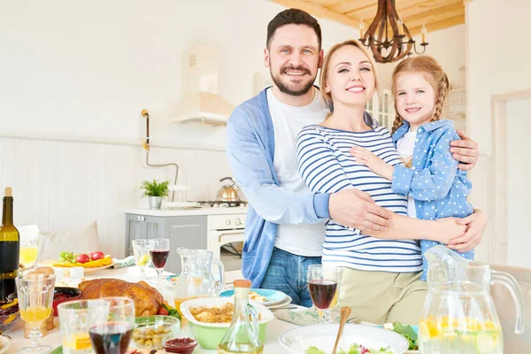 Portrait of happy young family standing at festive dinner table with delicious dishes and smiling at camera during  holiday  celebration in modern sunlit apartment, copy space