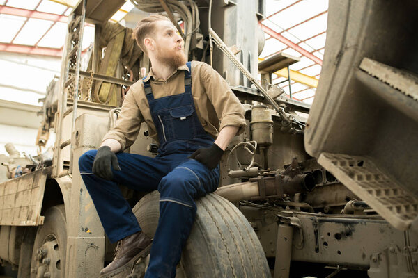 Young bearded male auto technician in overall sitting on wheel of big truck in repair shop and looking to the side