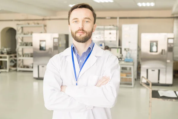 Serious confident successful young male engineer of pressure gauge plan wearing lab coat looking at camera and crossing arms on chest in workshop