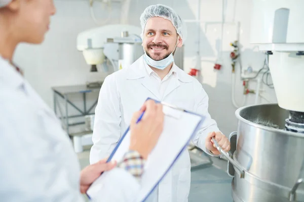 白衣の機械の近くに立って メモを作っている同僚を見て笑顔の製菓工場の従業員 — ストック写真