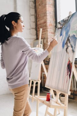 Yan görünümü portresi şövale Sanat stüdyo ayakta güneş ışığı altında resim resimler paleti tutan genç kadın sanatçı