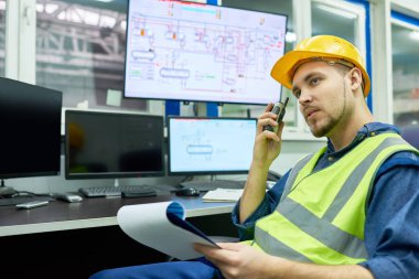Portrait of young man  wearing hardhat overseeing plant production process while sitting at desk in office and giving instructions by walkie-talkie, copy space clipart