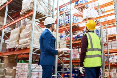 Ambar yöneticisinin ve mal ile uzun boylu raflar bakarak işçi ambar, stok envanter yapıyor kavganın görünüm portre geri