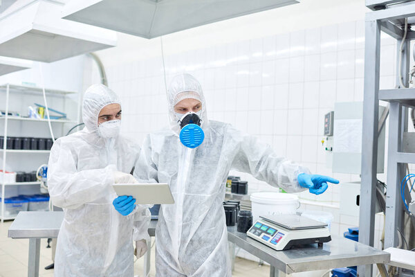 Sports nutrition production worker standing in protective clothing using tablet computer and indicating something to colleague.