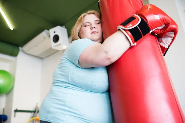 Low angle portrait of exhausted obese woman leaning on punching bag with eyes closed after extreme weight loss training in gym