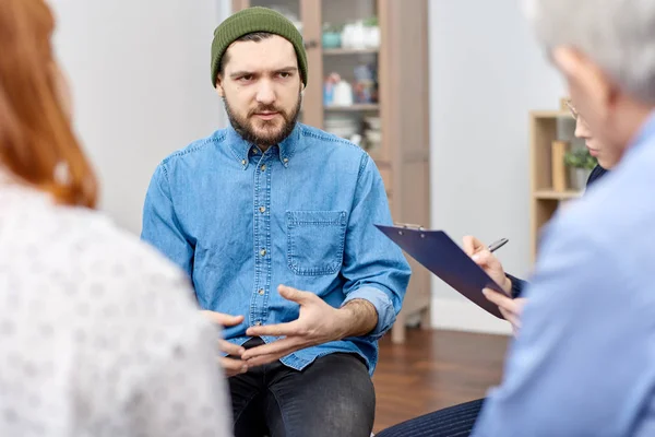 グループ療法のセッションに参加しながら高度なプロの心理学者および他の患者と彼の恐怖の議論デニム シャツを着た落ち込んでのひげを生やした男 — ストック写真
