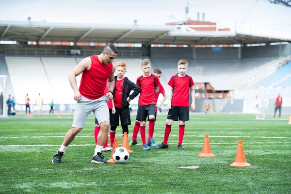 Full length portrait of junior football team practicing in stadium, focus on young coach leading ball in foreground, copy space