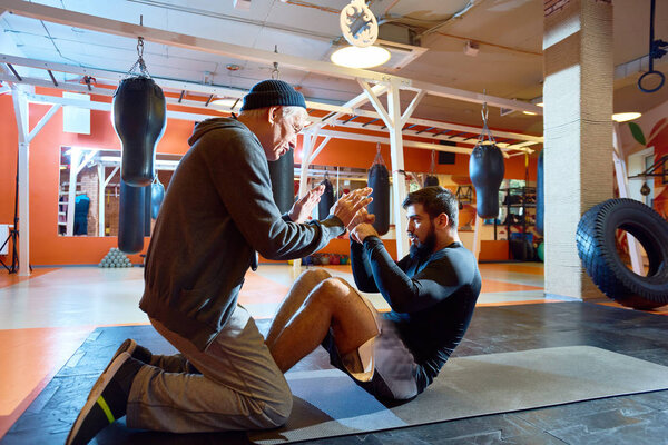 Старший мужчина обучение молодого боксера в тренажерном зале, помогая с абсом упражнения на полу и мотивации
. 