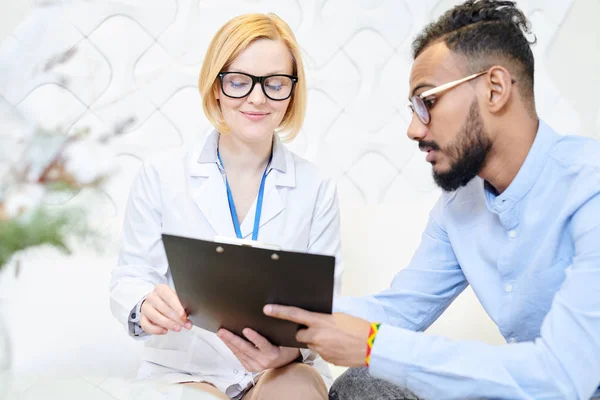 Profile view of bearded young patient holding clipboard in hands while filling on medical form, friendly doctor assisting him