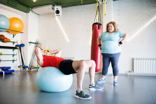 Full length portrait of obese young woman watching handsome man  doing exercises on fitness ball in gym