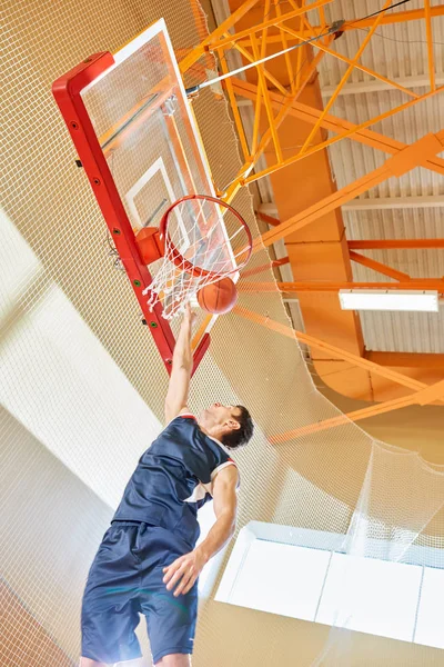 From below shot of young sportsman in basketball uniform trying to shoot goal while playing in gym.