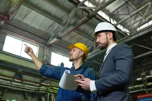 Side view portrait of two engineers discussing production in factory workshop, focus on businessman wearing hardhat and holding clipboard, copy space