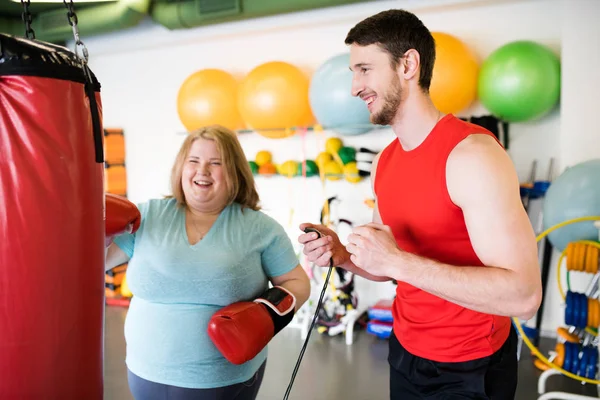 Portrait of happy obese woman hitting punching bag and laughing while exercising in gym with personal fitness coach