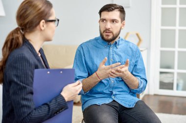 Handsome bearded patient having lack of social skills trying to solve problem with help of highly professional psychologist, interior of cozy office on background clipart