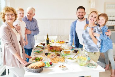 Portrait of happy two generation family enjoying dinner posing round festive table with delicious dishes and smiling at camera during holiday celebration in modern sunlit apartment, copy space clipart