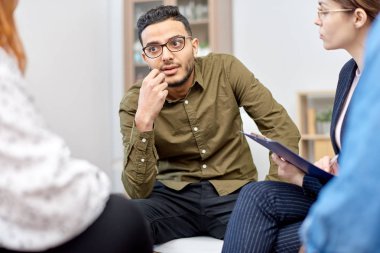 Portrait shot of handsome mixed-race man wearing eyeglasses participating in group therapy session at cozy office, friendly psychologist assisting her patients clipart