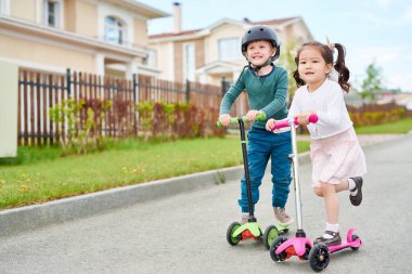 Tam uzunlukta portre sevimli iki sürme üç tekerlekli scooter çocuklar ve mutlu bir şekilde zengin yerleşim bölgesinde yolda giderken gülümseyen, uzay kopyalayın