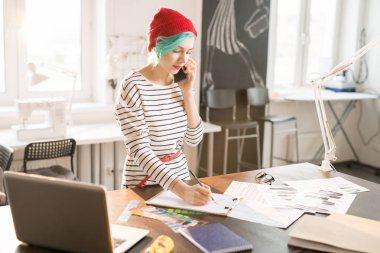 Çağdaş kadın moda telefonla konuşma ve güneşli atelier atölye çalışma masasında çizimler için özel yapılmış giysiler dururken çizim Tasarımcısı portresi