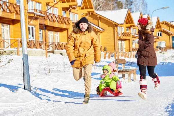 雪に覆われた道路に沿って実行していると 引っ張って若い白人カップルは町の明るい冬の日の幸せな小さな子供たちとそり — ストック写真