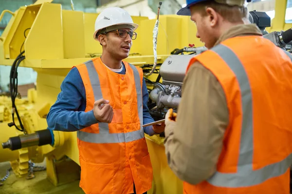 Two machine operators wearing reflective vests and protective helmets standing at production department of modern plant and trying to solve faced issue