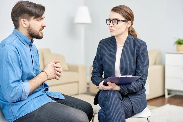 Handsome bearded man suffering from depression having consultation with highly professional psychiatrist at cozy office, she listening to him with concentration