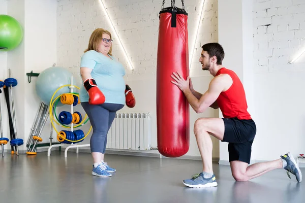 Full length portrait of obese woman in boxing training with fitness instructor in gym